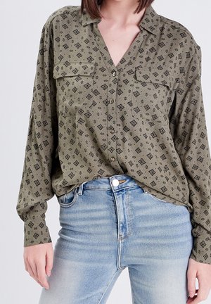 Femme portant une chemise boutonnée à motifs vert olive avec des poches poitrine et un jean taille haute bleu clair.