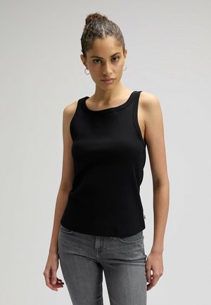 Kobieta z kręconymi włosami związanymi w kok, nosząca kolczyki koła, czarny, bezrękawnik i szare dżinsy na jasnoszarym tle.