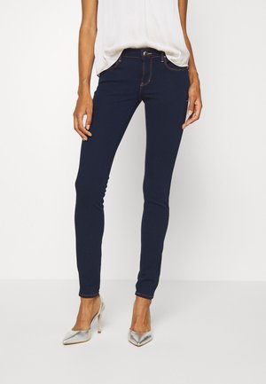 Vrouw draagt donkere blauwe strakke jeans, een los wit mouwloos topje en zilveren hoge hakken, staande tegen een effen witte achtergrond.