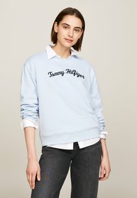 Felpa blu chiaro con scollo rotondo e logo ricamato "Tommy Hilfiger" in blu scuro. Indossata sopra una camicia bianca con colletto.