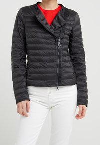 Doudoune noire avec fermeture éclair à l'avant, texture matelassée, coupe ajustée et col asymétrique. Associée à un pantalon blanc.