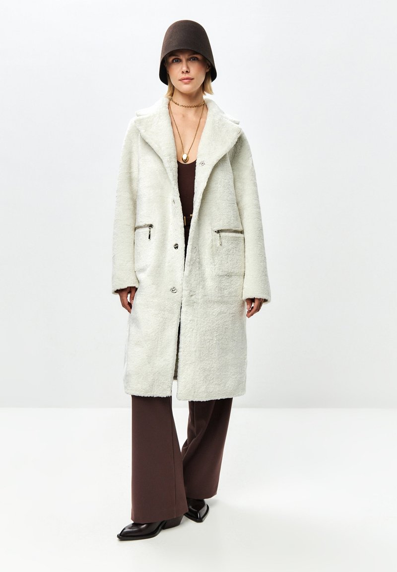 Donna che indossa un cappotto bianco texturizzato, pantaloni marroni a gamba larga, scarpe nere a punta, cappello marrone e collana d'oro in piedi davanti a uno sfondo neutro.