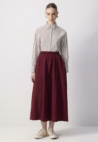 Chemise à rayures blanche et bleu marine, associée à une jupe bordeaux longueur cheville avec une ceinture élastique, et des baskets beiges.