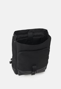 Sac à dos noir en tissu avec un fond en cuir lisse, doté d'une poche zippée à l'avant et de bretelles réglables. L'intérieur est équipé de compartiments organisés.