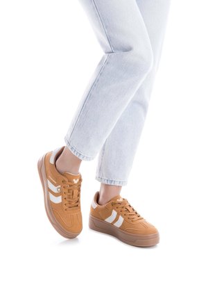 Zapatillas de ante beige con rayas blancas, diseño de cordones, suelas de goma y cuello acolchado. Llevadas con jeans azules claros con dobladillo.