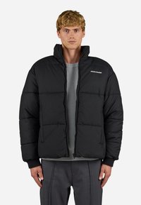 Jachetă puf de culoare neagră, cu fermoar frontal, formă boxy, manșete ribbate și logo-ul "Pegador". Poartă peste un pulover texturat gri și pantaloni gri.