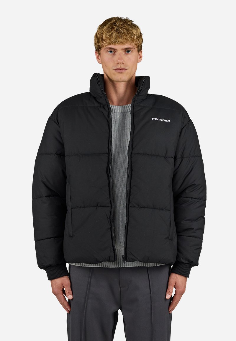 Jachetă puf de culoare neagră, cu fermoar frontal, formă boxy, manșete ribbate și logo-ul "Pegador". Poartă peste un pulover texturat gri și pantaloni gri.