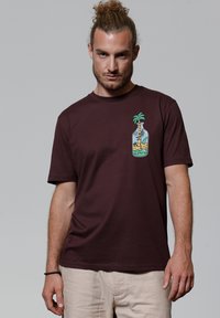Bordeauxfarbenes Baumwoll-T-Shirt mit kurzen Ärmeln, das auf der linken Brustseite ein Grafikbild eines Palmenbaums in einer Flasche zeigt. Glatte Textur.