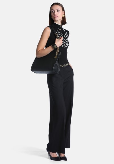 Femme portant un haut sans manches noir avec motif blanc, pantalon noir, talons noirs, boucles d'oreilles dorées, bracelet, et tenant un grand sac à main noir.