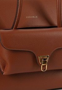 Bruine leren handtas met een gestructureerd oppervlak en een gouden metalen sluiting. Heeft afgeronde hoeken en een zichtbaar logo vlak bij de bovenkant. Dubbel handvat ontwerp.