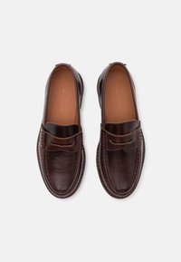 Barna bőr slip-on cipők sima felületűek, kerek orrúak, és kontrasztos varrással rendelkeznek. Díszes sáv található az elején.