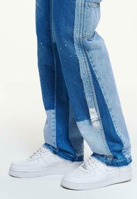 Kojos dėvint dvispalvius mėlynos spalvos džinsus su dažų purslais ir baltus Nike Air Force 1 sportinius batelius ant balto fono.