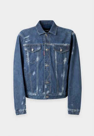 Jeune homme portant une veste en jean usée et un jean, un pull col en V couleur rouille, des bottes marron clair, une casquette noire avec logo et une ceinture blanche de marque visible.