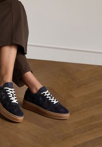 Marineblaue Wildleder-Sneaker mit weißen Schnürsenkeln und einer Gummisohle in Tan. Mit abgerundeten Designelementen und einem gepolsterten Absatz für zusätzlichen Komfort.