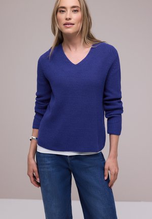 Kvinde iført en struktureret blå V-hals sweater over en hvid skjorte med blå jeans, stående foran en ensfarvet lysegrå baggrund.