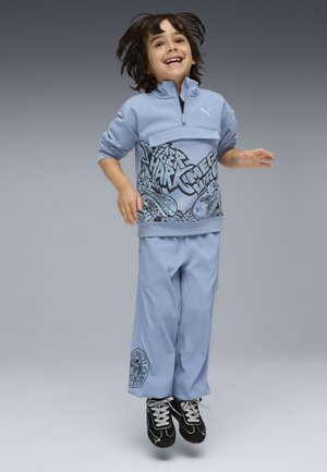Enfant souriant sautant dans les airs, portant un sweat-shirt graphique bleu clair et un pantalon assorti, avec des baskets noires sur un fond dégradé gris.