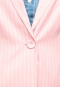 Blazer rose clair à fines rayures boutonné sur une chemise en denim bleu avec un bouton blanc visible au col.