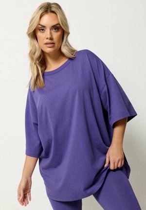 Femme blonde portant un t-shirt violet ample surdimensionné et des leggings assortis, posant devant un fond clair uni.