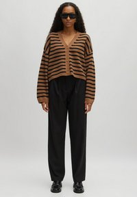 Bruine en zwarte gestreepte korte cardigan met drie knopen, gecombineerd met losse zwarte broeken en zwarte schoenen, tegen een neutrale achtergrond.
