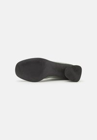 Suela de un zapato verde con suela de goma negra que presenta un diseño de huellas con patrón de hojas, mostrado sobre un fondo blanco liso.