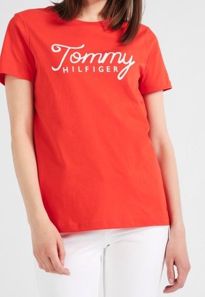 Kobieta ubrana w jasnoczerwoną koszulkę z krótkim rękawem z logo "Tommy Hilfiger" oraz białe spodnie na jednolitym tle.