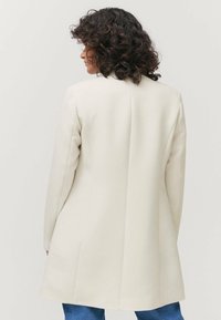 Abrigo beige ajustado con una silueta recta, mangas largas y líneas limpias. Tejido suave con un detalle de abertura trasera para facilitar el movimiento.