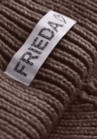 Bruine gebreide stof met een verhoogde textuur; bevat een wit label met "FRIEDA" in zwarte tekst.
