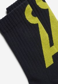 Schwarze Socken mit einem großen gelben Design, hergestellt aus einem strukturierten Strickmaterial. Das Muster enthält kräftige, stilisierte Schriftzüge.