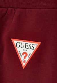 Burgundy puuvillakangas, jossa on triangelimainen Guess-logo-tilausteksti punaisena, valkoisena ja mustana sekä teksti "WASHED JEANS U.S.A." ja kysymysmerkki.