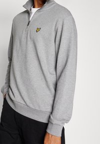 Felpa grigia con cerniera realizzata in tessuto morbido. Presenta un colletto alto, polsini a coste e un piccolo emblema giallo sul petto.