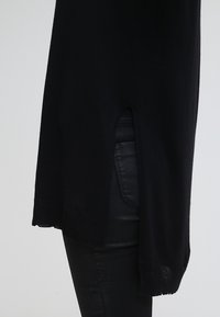 Un long pull noir avec une fente sur le côté porté sur un pantalon en cuir noir sur fond clair.