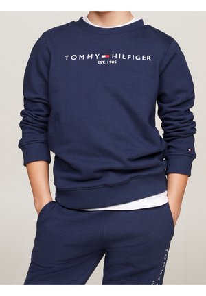 Granatowy komplet dresowy Tommy Hilfiger z logo i napisem „Est. 1985,” ręce w kieszeniach, widoczna biała koszula pod spodem.