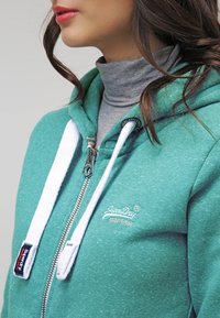 Sudadera con capucha en color azul verdoso, con cordones blancos, cierre de cremallera y logo bordado. Lleva una capa de debajo en cuello alto gris. Tejido suave y texturizado.