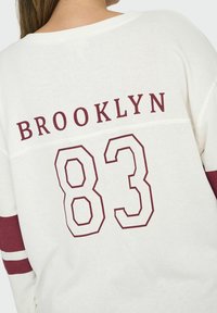 Bílá mikina s vínovými akcenty, na které je nad číslem "83" napsáno "BROOKLYN". Má pruhované rukávy a měkkou, strukturovanou tkaninu.