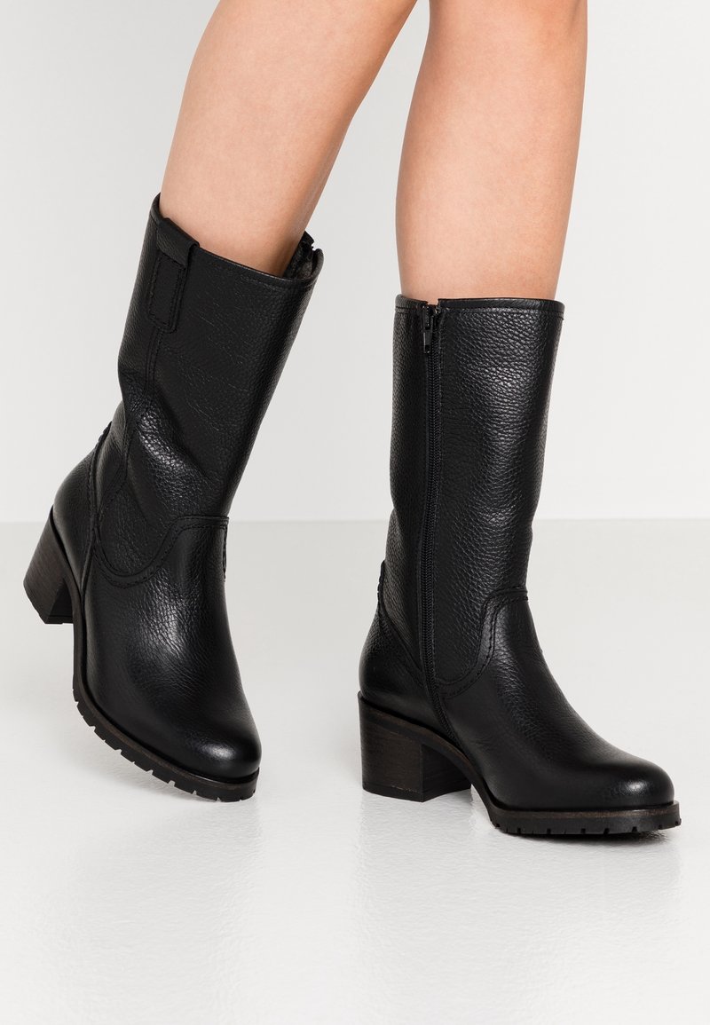 Botas de cuero negras a la mitad de la pantorrilla con cremalleras laterales y tacones cuadrados, llevadas sobre piernas desnudas contra un fondo blanco liso.
