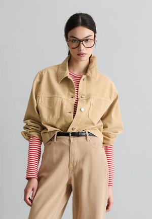 Młoda kobieta w oversize'owej beżowej kurtce, czerwono-białej w paski koszuli z długim rękawem, beżowych spodniach, czarnym pasku i dużych okularach w oprawkach w kolorze żółwia.