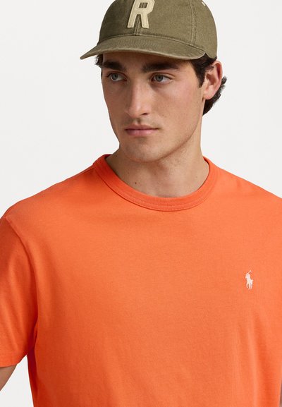 Orange bomullströja med rund halsringning och korta ärmar, med en liten, vit broderad logotyp på vänster bröst.