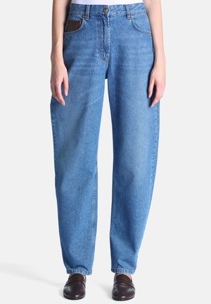 Žena v modrých džínsoch s vysokým pásom a voľným strihom a tmavohnedých slip-on topánkach, stojaca pred jednofarebným svetlým pozadím.