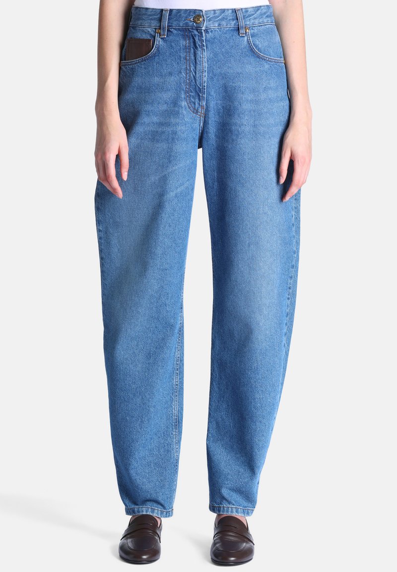 Femme portant un jean bleu à taille haute et coupe décontractée, ainsi que des chaussures marron foncé sans lacets, debout devant un fond clair uni.