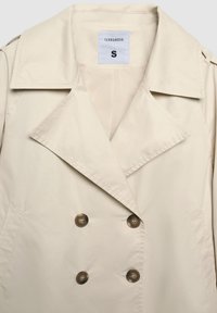 Trench-coat beige à double boutonnage avec de larges revers, quatre boutons, des poches latérales et une étiquette taille small à l'intérieur du col.