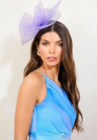Abito azzurro chiaro con un design drappeggiato, scollo all'americana e dettagli in catena dorata. Il fascinator in rete viola aggiunge una texture a strati e volume.