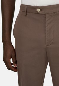 Personne à la peau foncée portant un pantalon marron sur mesure avec passants de ceinture et bouton, main relâchée le long du corps sur un fond blanc.