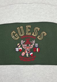 Zelená a šedá mikina s nápisem "GUESS ORIGINALS" tučně. Centrální vyšívaný design obsahuje jelena a různé heraldické štíty.