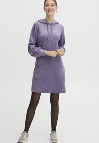 Lila velour hoodieklänning med långa ärmar, justerbar dragsko och en liten logodetalj vid fållen. Kombineras med svarta tights och sneakers.