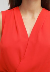Red sleeveless top with a deep V-neckline, made of smooth fabric, featuring draped detailing at the chest area.