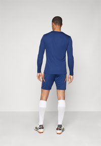 Maglietta sportiva a maniche lunghe blu della marina e pantaloncini abbinati con dettagli arancioni, accompagnati da calzini bianchi e scarpini da calcio neri/grigi.