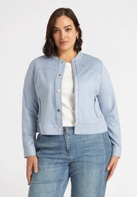 Chaqueta de cuero sintético azul claro con cuello redondo, bolsillos con cremallera y herrajes plateados, combinada con una blusa blanca y jeans de mezclilla.