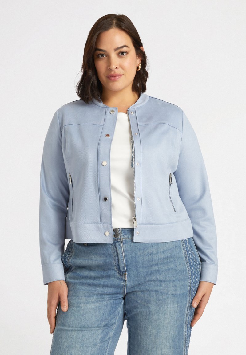 Chaqueta de cuero sintético azul claro con cuello redondo, bolsillos con cremallera y herrajes plateados, combinada con una blusa blanca y jeans de mezclilla.
