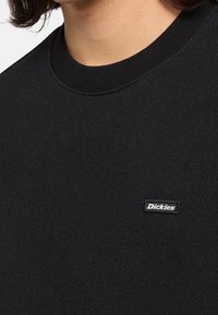Gros plan d'une personne portant un sweat-shirt noir à col rond avec une petite étiquette rectangulaire du logo Dickies sur la poitrine.