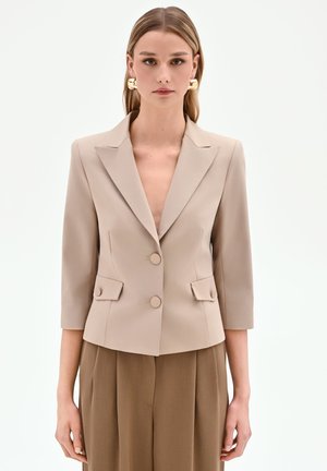 Donna che indossa un blazer beige su misura con maniche a tre quarti e due bottoni frontali, abbinato a pantaloni marroni a vita alta.